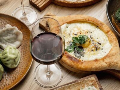 A spread of foods on a wooden table: a cheese-filled bread bowl, a wooden board with grilled meat and onions, dumplings with sauces, a glass of red wine, and a burger in the background.