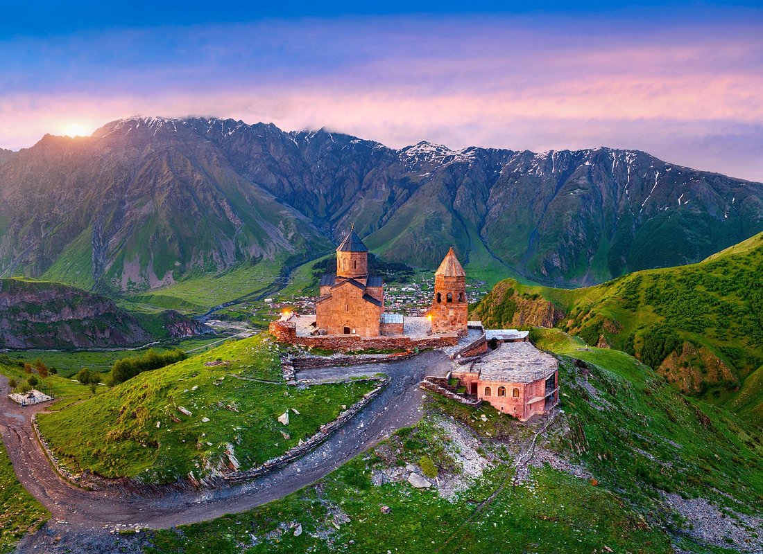 Stone church and twin towers atop a hill overlooking a green valley, with distant snow-capped mountains at sunset.