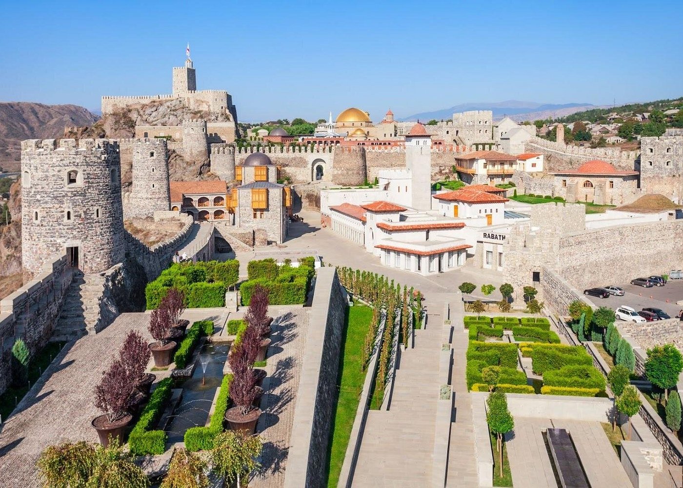 Aerial view of Rabath Fortress: stone walls and towers surround a formal garden with trimmed hedges and pathways under a clear blue sky.