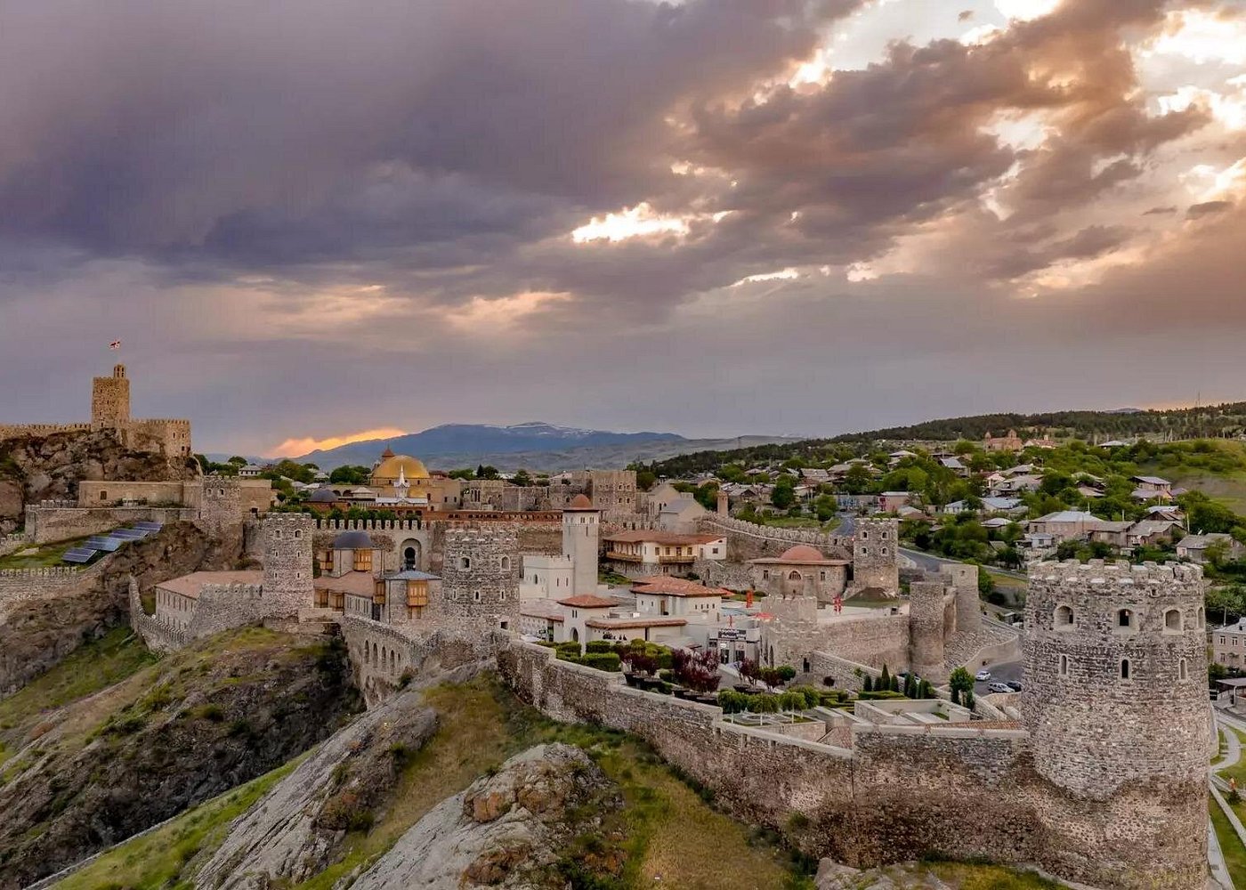 Medieval fortress city with stone walls and towers atop a rocky hill at sunset.