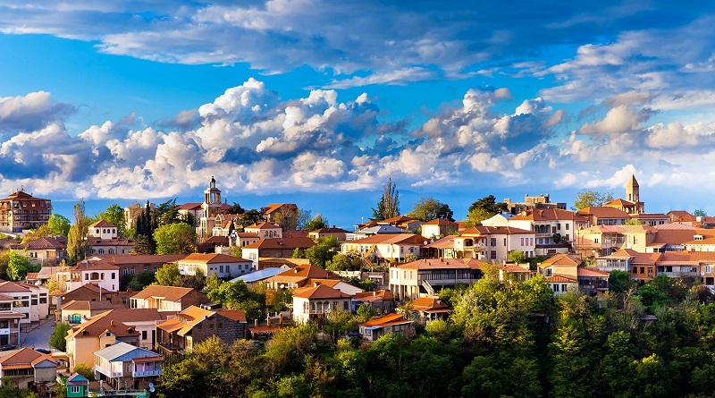 Hillside town with colorful houses and trees under a bright blue sky with fluffy clouds.