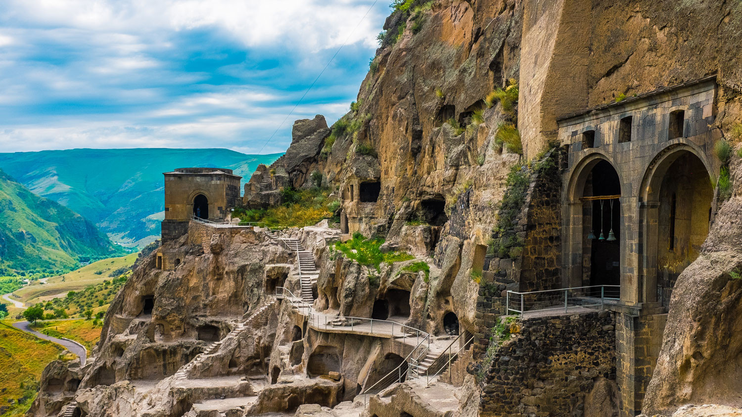 Rock-cut monastery carved into a cliff with arched entrances and stairways, overlooking a green valley.
