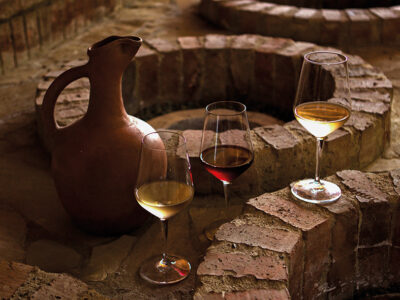 A rustic clay jug surrounded by four wine glasses of white, red, and pale white wine on a weathered brick circular surface.