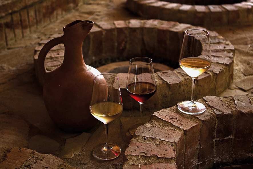 A rustic clay jug surrounded by four wine glasses of white, red, and pale white wine on a weathered brick circular surface.